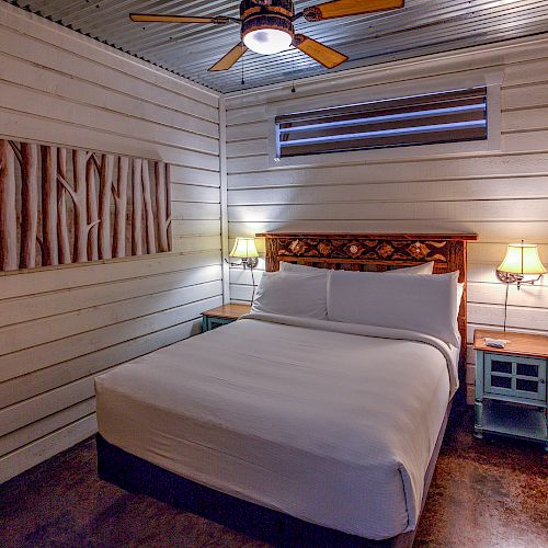 This image shows a cozy bedroom with a double bed, two nightstands, two lamps, a ceiling fan, and wall decor. The room has wooden walls and flooring.