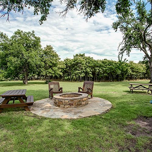 A grassy outdoor area with a stone fire pit surrounded by wooden chairs, picnic tables, and a swing, with trees in the background.