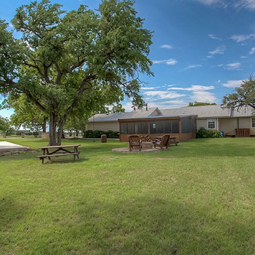 A spacious yard with a house in the background, featuring trees, a picnic table, and an outdoor seating area with a fire pit under a clear sky.