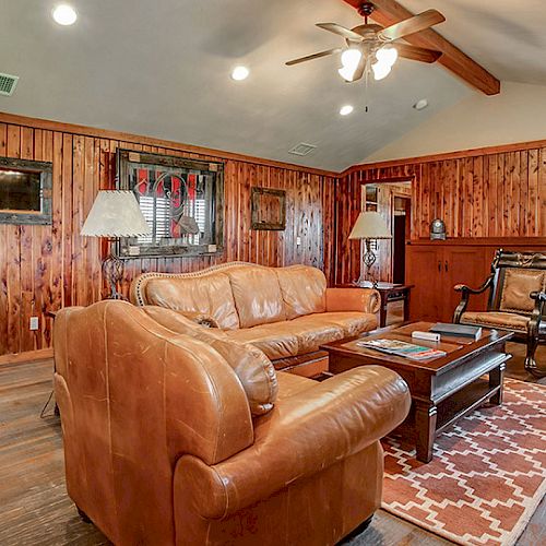 A cozy, wood-paneled living room with a leather sofa and armchairs, coffee table, area rug, ceiling fan, and framed wall decorations.