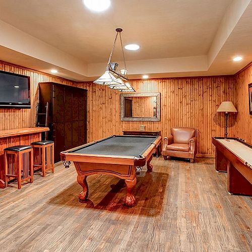 A cozy game room with wood paneling, featuring a pool table, shuffleboard, TV, bar area with stools, leather chair, lamp, and wall art.