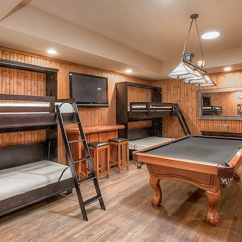 The image shows a cozy room with wooden walls featuring bunk beds, a pool table, a TV, bar stools, a mirror, and an armchair.