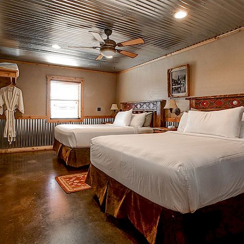 A cozy rustic bedroom with two queen beds, robes hanging, a window, wooden and corrugated metal walls, and a ceiling fan.