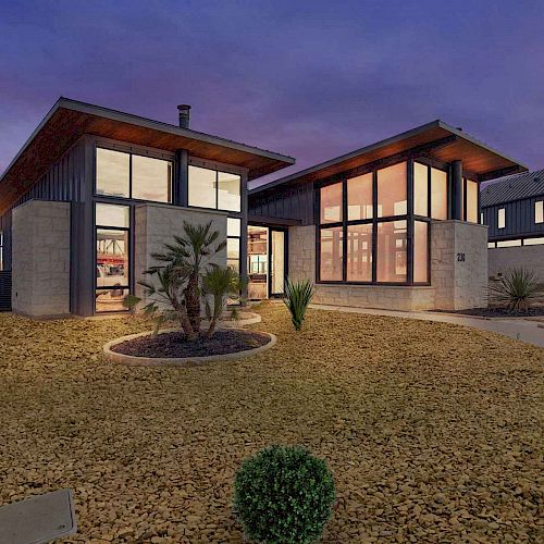 A modern, single-story house with large windows, surrounded by a landscaped yard with minimalistic plants and pathways at sunset.