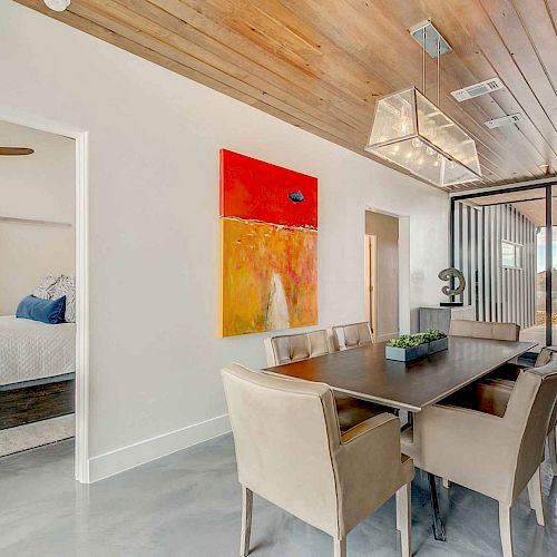 A modern dining room features a wooden ceiling, contemporary light fixtures, abstract art, six beige chairs, and a dark table adjacent to a bedroom.