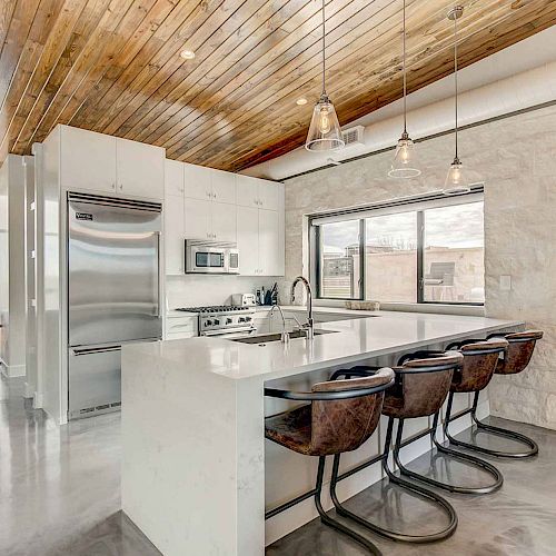 A modern kitchen with wooden ceiling, marble island, stainless steel appliances, pendant lights, and four bar stools, leading to a living area with a window.