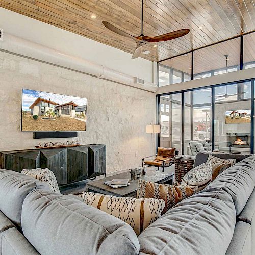 A modern living room features a sectional sofa, wall-mounted TV, wooden ceiling, stone wall, and glass doors leading to an outdoor seating area with a fireplace.