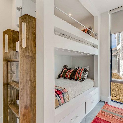 This image shows a compact bedroom with a set of built-in bunk beds, colorful pillows and blankets, a small wooden shelf, and a window with a view outside.