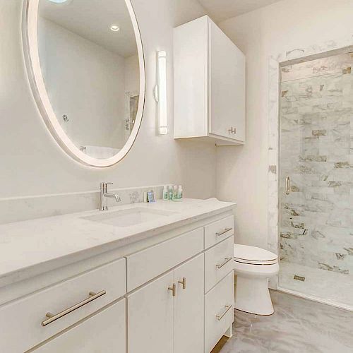 A modern bathroom with white cabinets, an oval mirror, a toilet, and a walk-in shower with a glass door, all in a bright neutral color scheme.