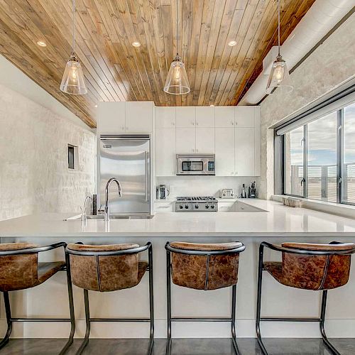 A modern kitchen with wooden ceiling, hanging lights, a large window, bar stools, white countertops, sink, refrigerator, oven, and microwave.