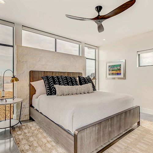 A modern bedroom with a wooden bed, white bedding, black patterned pillows, side table with a lamp, large windows and a ceiling fan ends the sentence.