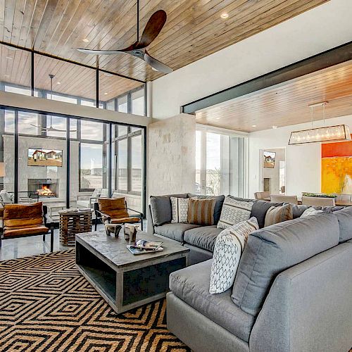A modern living space with large windows, wooden ceiling, a fan, gray sectional sofa, patterned rug, and contemporary art on the dining area wall.