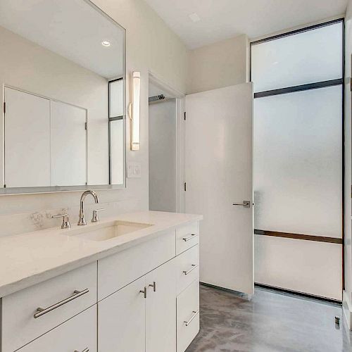 The image shows a modern bathroom with a large mirror, a sink with a countertop, cabinets, a frosted glass door, and a glass-enclosed shower.