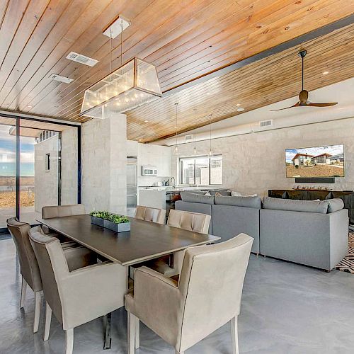 A modern living and dining area with a wooden ceiling, large dining table, chairs, sofas, TV, and large windows overlooking an outdoor view.