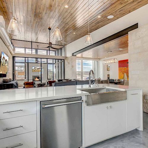 A modern kitchen features a stainless steel dishwasher, a large sink, white cabinetry, pendant lighting, and a wooden ceiling.