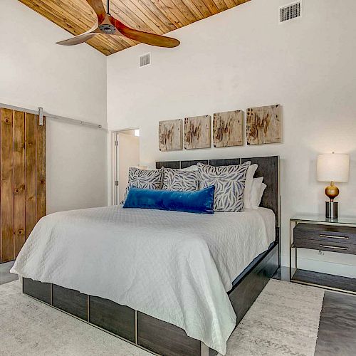A modern bedroom features a wooden ceiling fan, a bed with white and blue bedding, a side table with a lamp, and a sliding barn door.