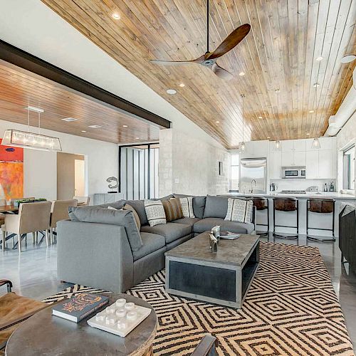 A modern open-concept living room and kitchen with wooden ceilings, a gray sectional sofa, a geometric rug, and dining area with bright wall art.