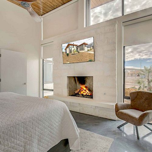 A modern bedroom features a fireplace, flat-screen TV, single chair, large windows, and a neatly made bed with white linens and blue pillows.