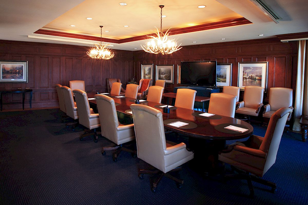 A conference room with a long table, leather chairs, wall art, two chandeliers, and a large screen on the wall; set up for a meeting with notepads.