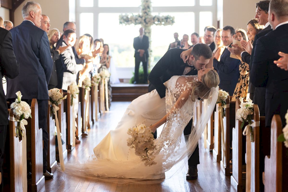A couple kisses in a wedding ceremony while guests watch, captured in a church aisle adorned with flowers, in front of a windowed backdrop.