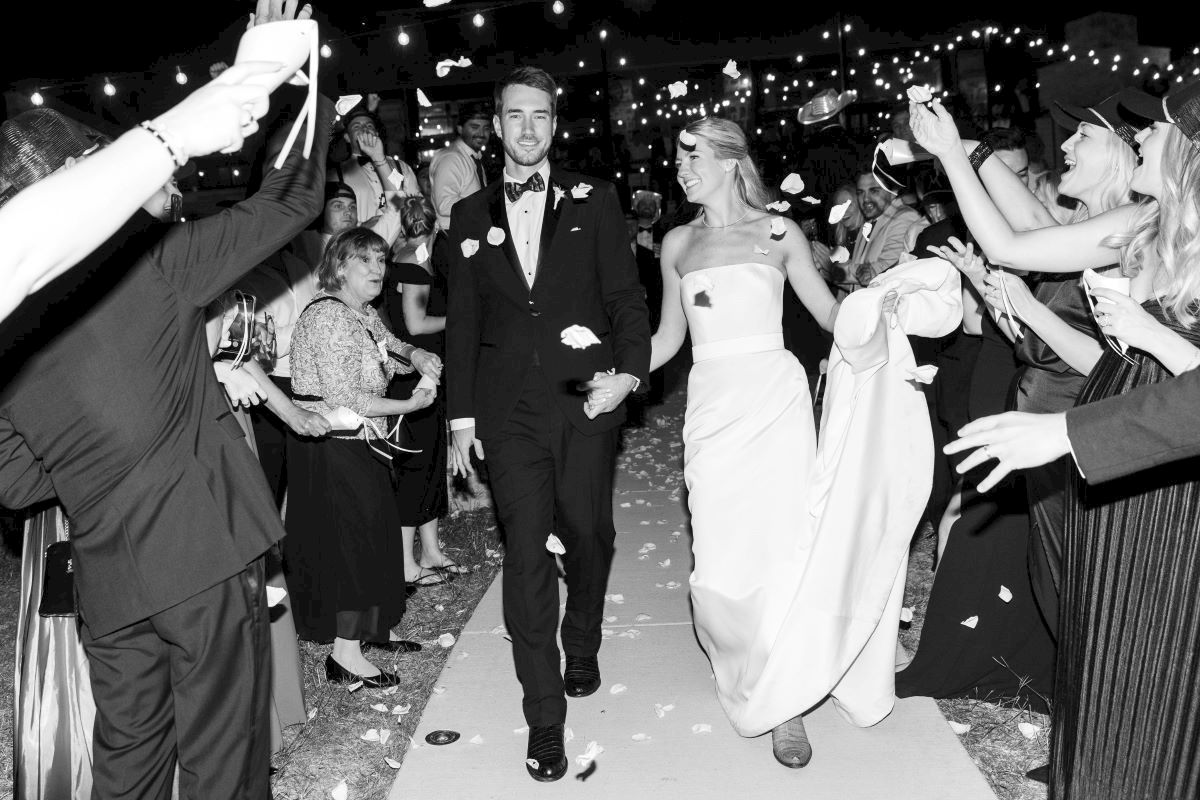 A newlywed couple walks down an aisle, surrounded by celebrating guests throwing petals at night, under string lights.