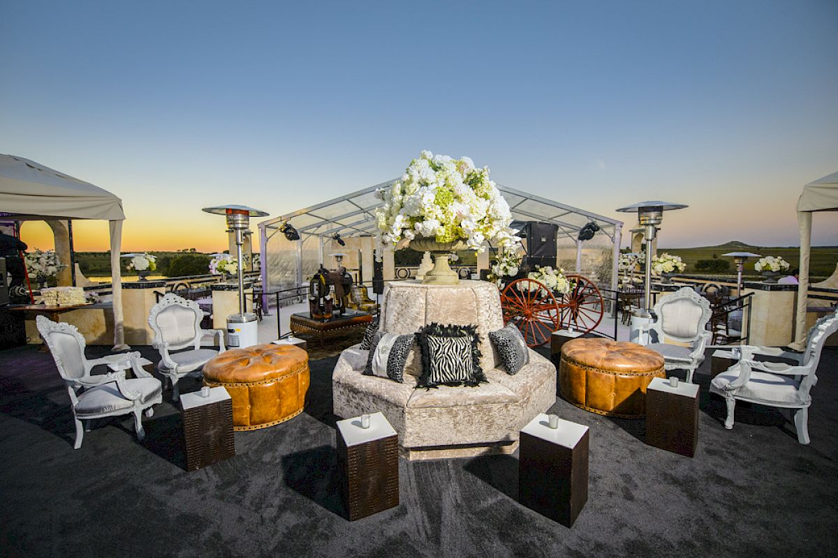 An outdoor event setup with elegant seating, plush furniture, decorative flowers, and tents under a sunset sky.