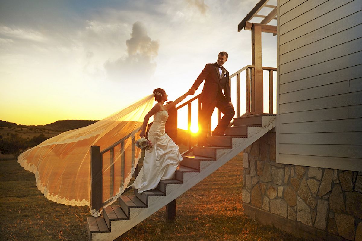 A bride and groom descend outdoor stairs at sunset, with the bride's veil flowing in the wind, capturing a picturesque moment.
