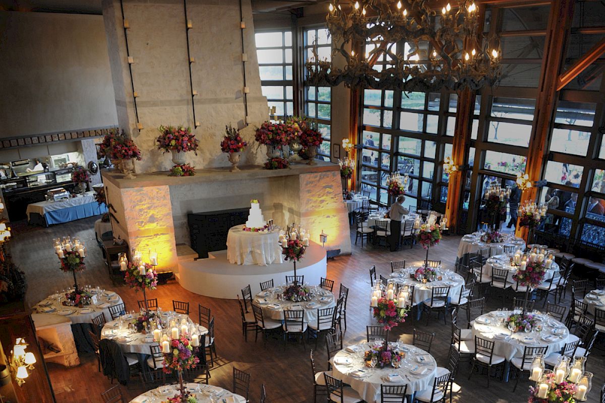 A decorated event space with round tables set for a reception, featuring floral arrangements and a central white tiered cake on display.