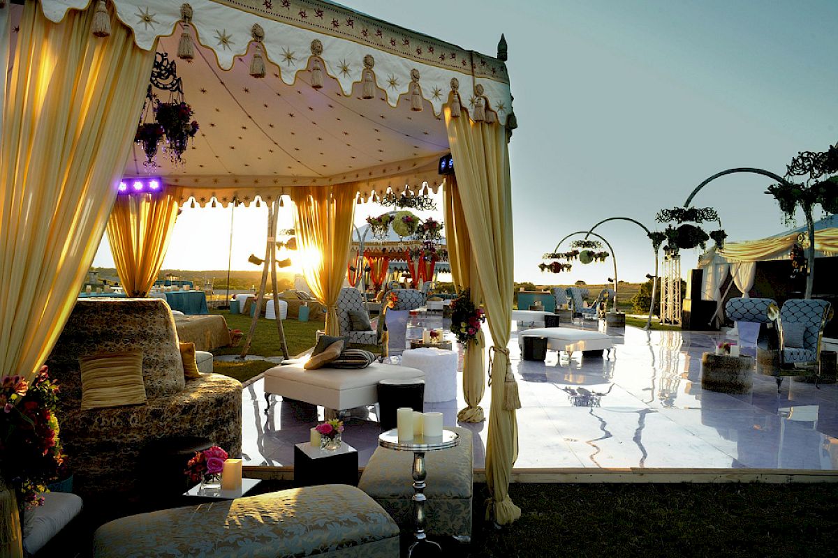 The image shows an elegant outdoor event setup with draped tents, luxurious seating, decorated tables, and lighting fixtures during sunset.