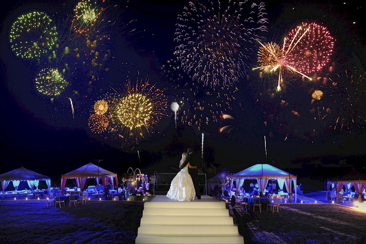 A bride in a white dress stands on steps, watching colorful fireworks in the night sky, with lit tents and guests in the background.