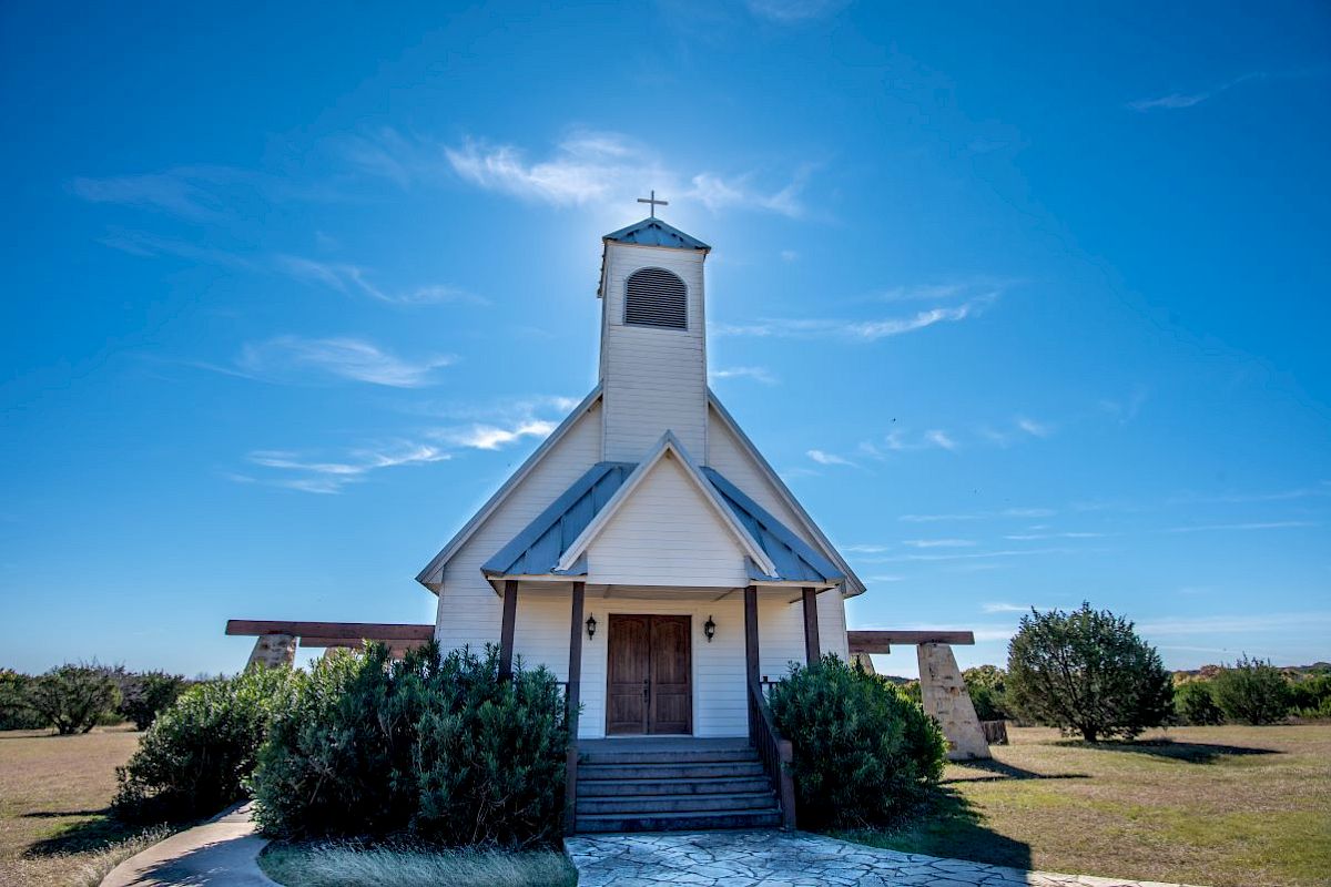 A small white church with a bell tower under a clear blue sky, surrounded by greenery and open fields, stands peacefully in the sunlight.