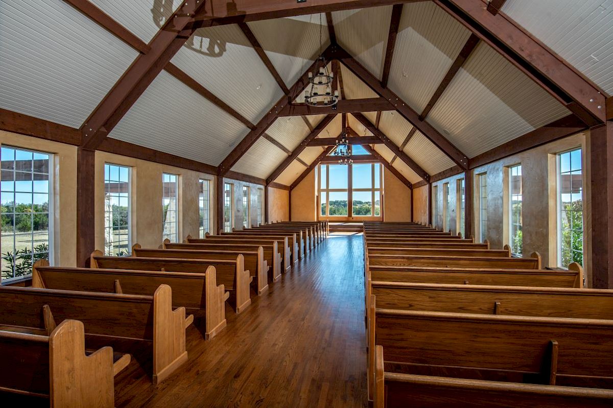 The image shows a wooden chapel interior with rows of pews, large windows, and a high ceiling, leading to a bright view outside.