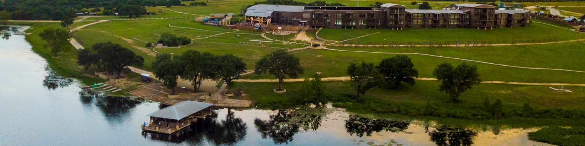 A large lakeside property with buildings, trees, and a dock over the water, set in a vast green landscape under a slightly cloudy sky.
