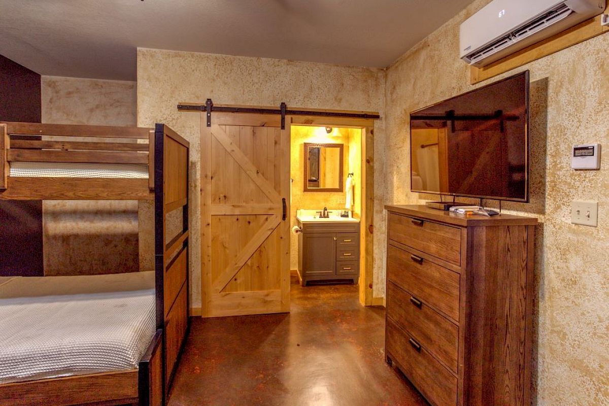 The image shows a bedroom with bunk beds, a dresser with a TV, and a bathroom with a sliding barn door open, revealing a sink.