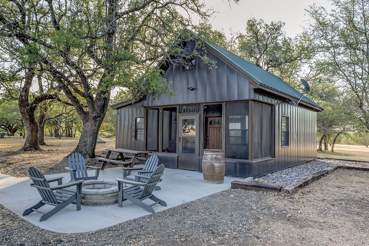 A small cabin with a metal roof surrounded by trees features outdoor seating and a fire pit area.