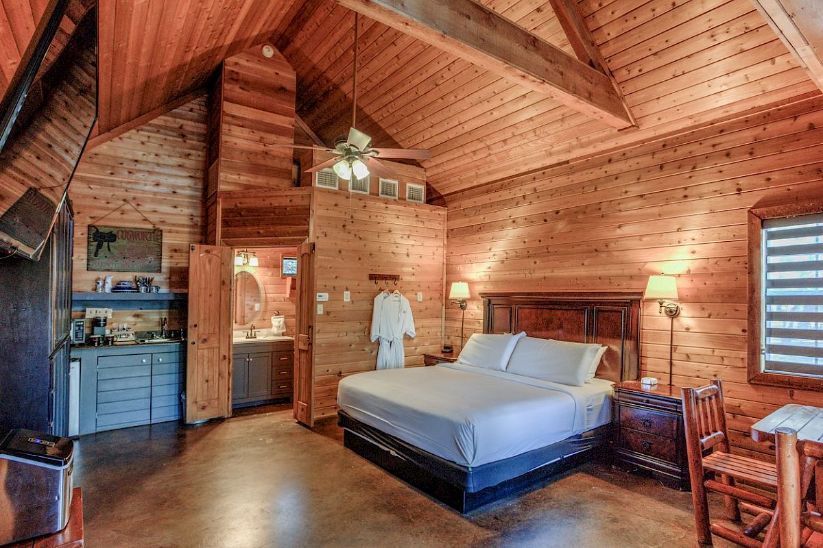A cozy cabin room with wooden walls, a large bed, kitchenette, and a chair, featuring rustic decor and high ceilings, creating a warm atmosphere.