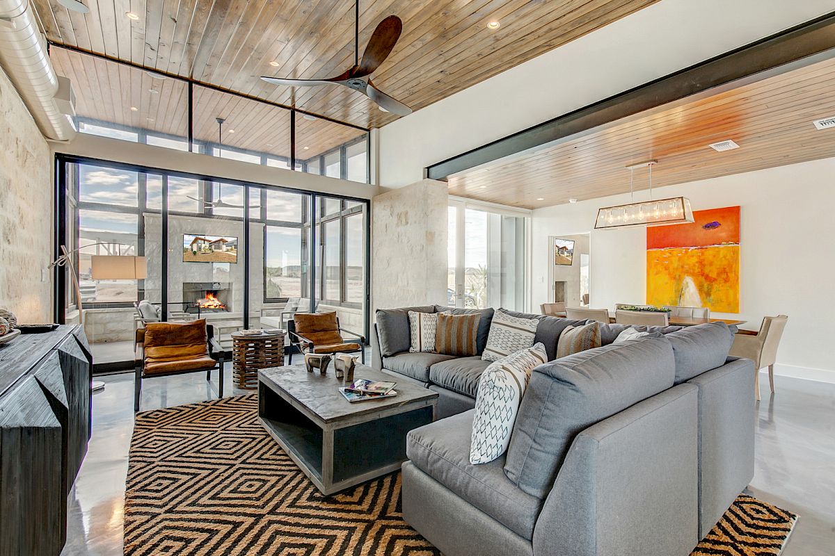 A modern living room with a gray sectional sofa, geometric rug, fireplace, and large windows. A vibrant painting hangs near the dining area.