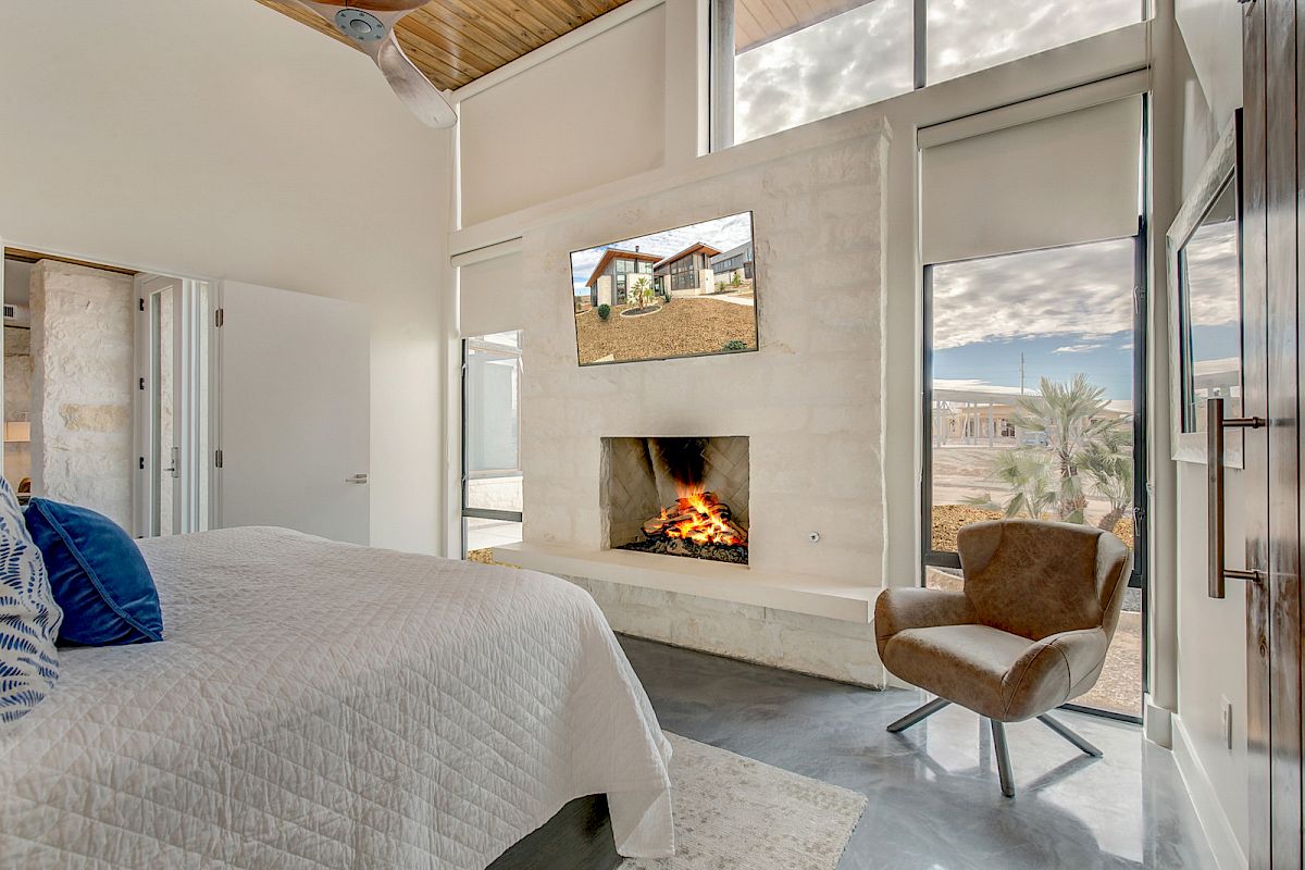A cozy bedroom with a bed, armchair, fireplace, and large windows, featuring a landscape on a TV above the fireplace.