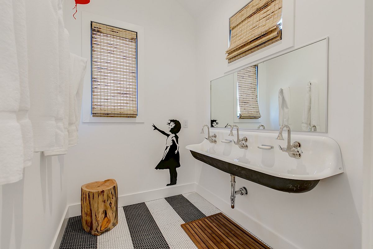 A bathroom with a long sink, wall art of a girl, wooden stool, striped floor tiles, and window shades.