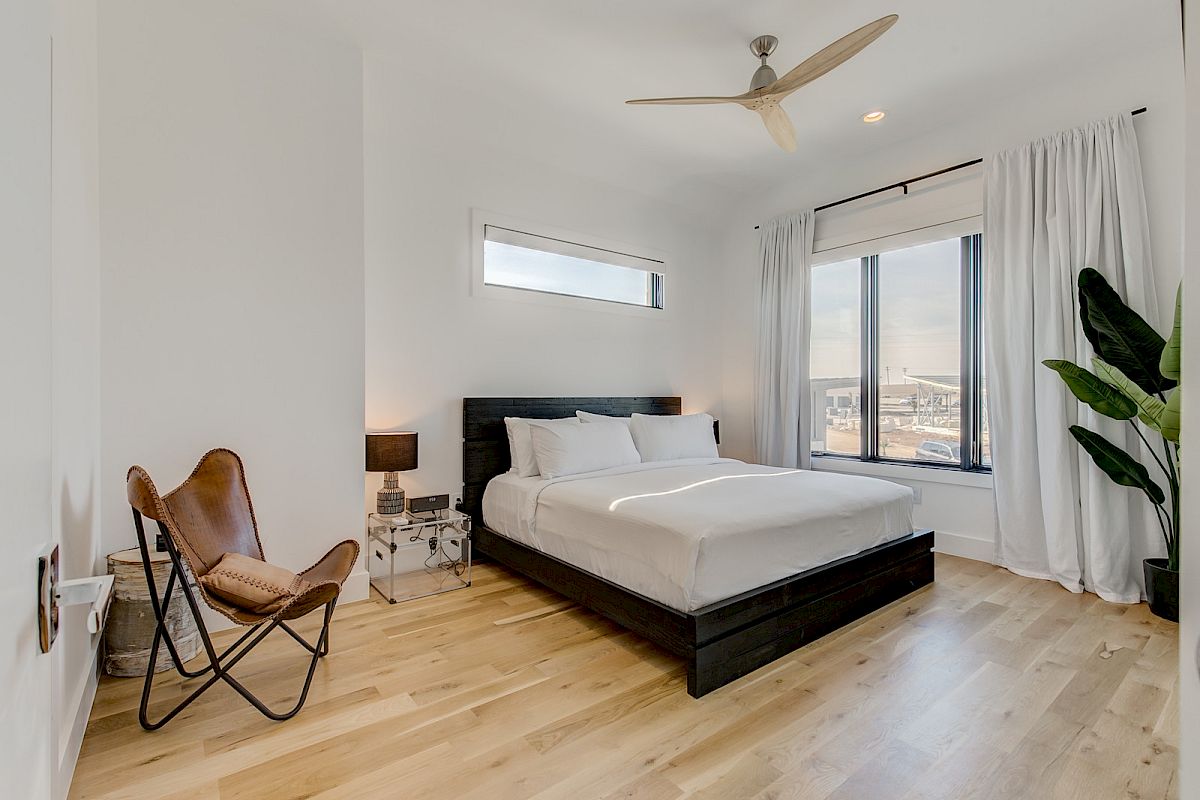 A modern bedroom with a bed, two windows, a wooden floor, a chair, lamp, plant, and ceiling fan.