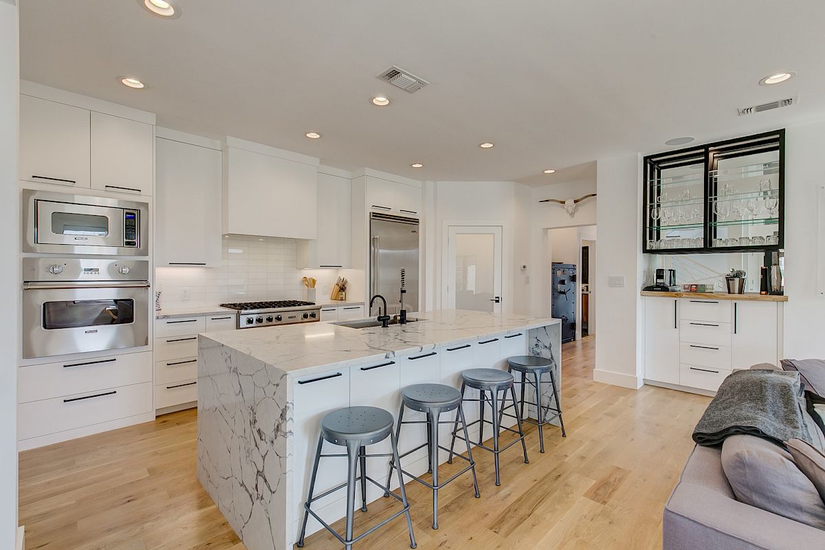 A modern kitchen with marble island, bar stools, stainless steel appliances, light wood flooring, and shelves with glasses.