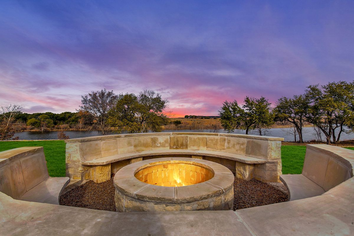 A cozy outdoor fire pit surrounded by stone seating overlooks a peaceful lake, with a colorful sunset sky in the background.