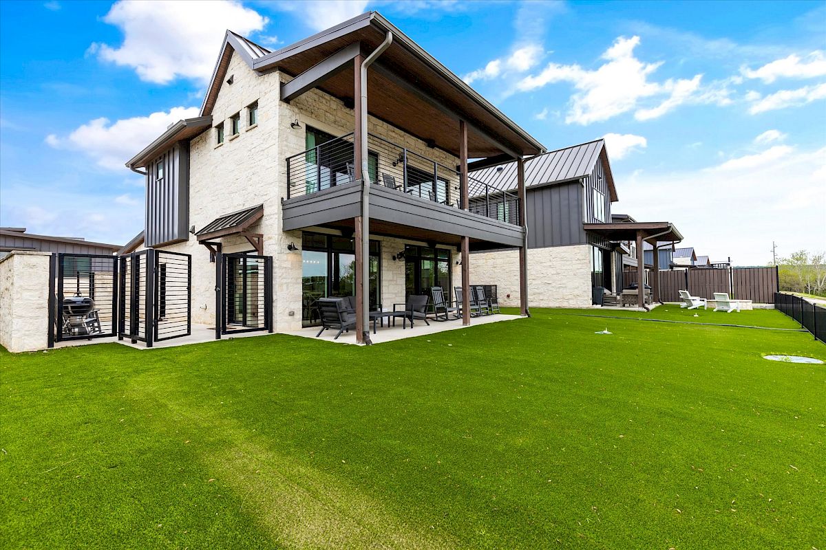 A modern two-story house with a stone exterior, large windows, and green lawn. There's a balcony and patio seating.
