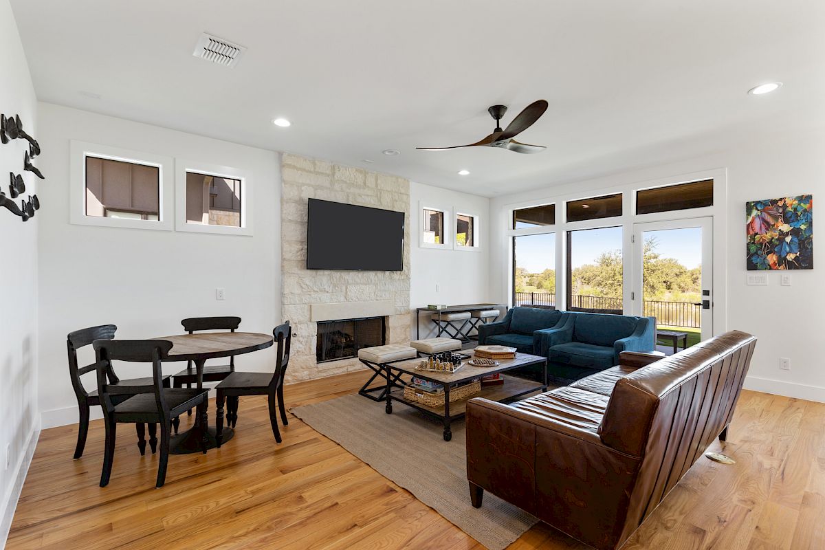A modern living room features a dining table, seating area, stone fireplace, wall-mounted TV, hardwood floors, and large windows.