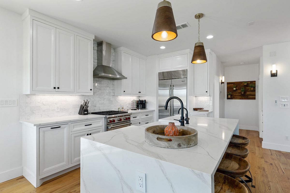 A modern kitchen with white cabinets, marble island, pendant lights, stainless steel appliances, and wooden barstools.