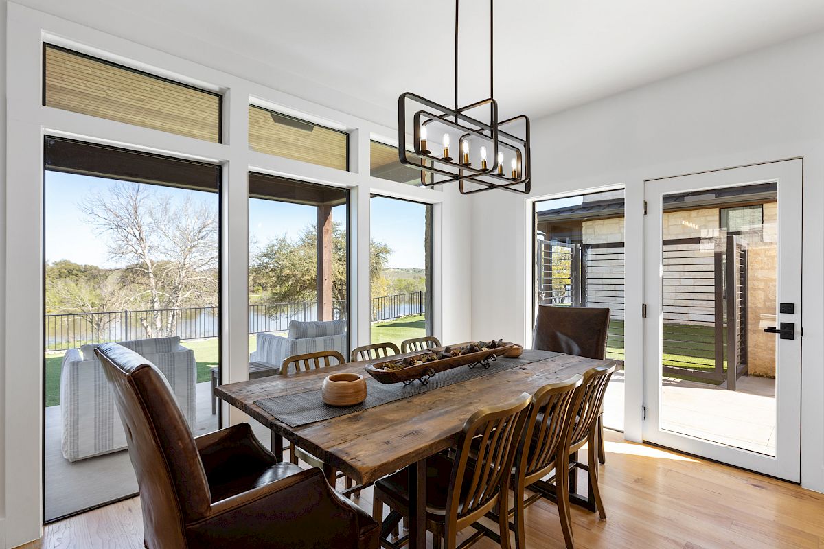 A dining room with a wooden table, several chairs, large windows, and a view of an outdoor patio is shown.