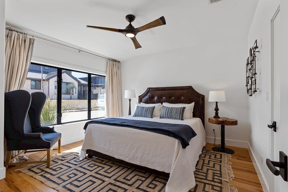 A modern bedroom with a bed, two lamps, a large window, an armchair, and a ceiling fan, featuring minimalist decor.