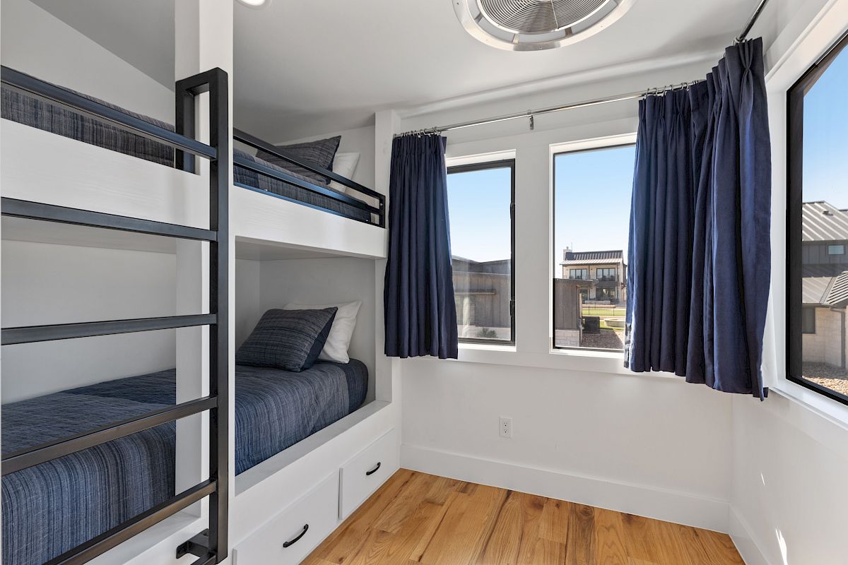 The image shows a small bedroom with bunk beds, blue curtains, white walls, and windows providing natural light.
