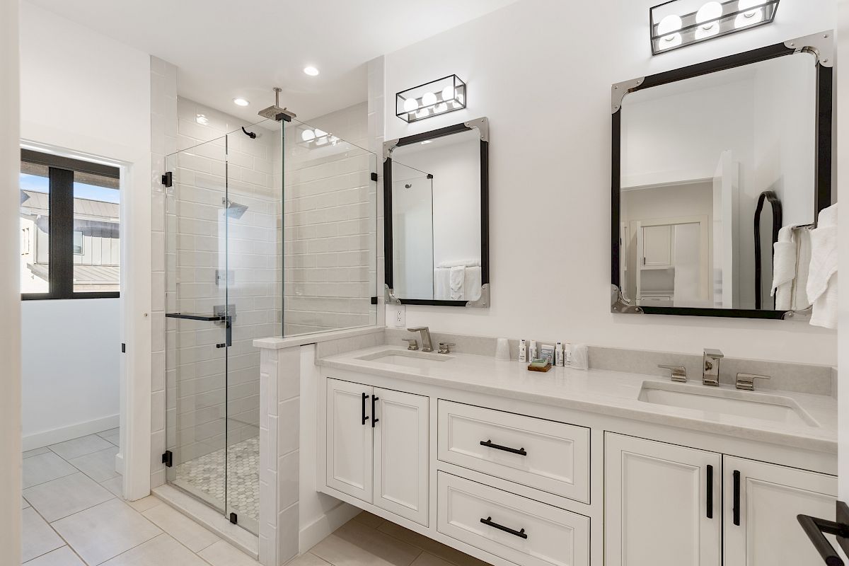 A modern bathroom with a walk-in shower, dual sinks, and large mirrors. Bright lighting and a minimalistic design enhance the space.