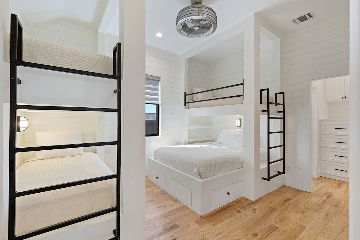 The image shows a modern bedroom with white shiplap walls, bunk beds, ladders, and wooden flooring, featuring built-in storage drawers.
